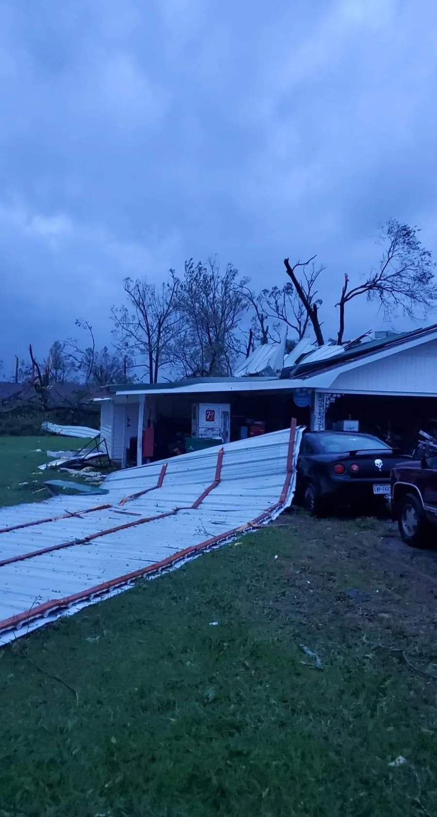 Metal roof panels torn off a house and carport by straight-line wind, lying across the lawn under a dark storm sky
